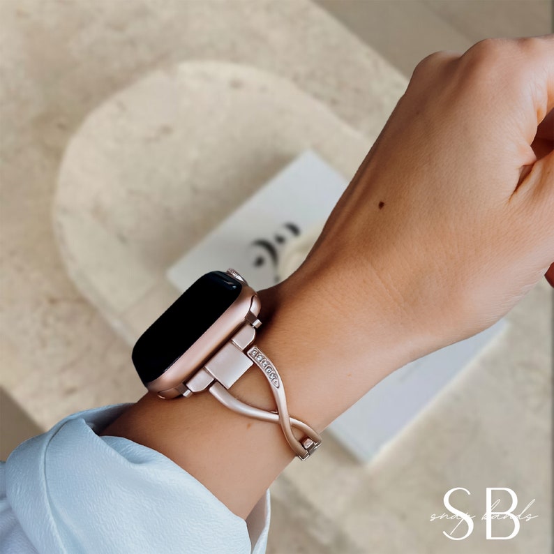 A rose gold Apple Watch with a rose gold band featuring a criss-cross design and small clear stones. The band is engraved with the word "SELENA".