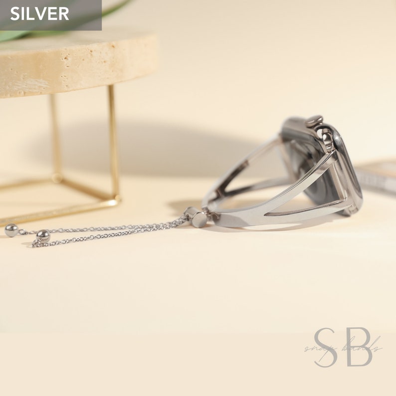 Silver watch band with a chain and beads. The band is designed to fit a square-shaped watch face. The band is made of metal and has a sleek, modern design.