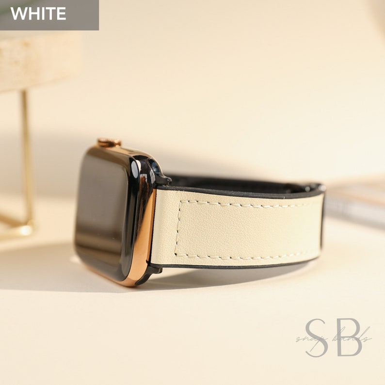 Cream-colored Apple Watch band with a black interior and gold-tone watch face. The band is made of a smooth material and features white stitching. The word "WHITE" is in a white box in the upper left corner.