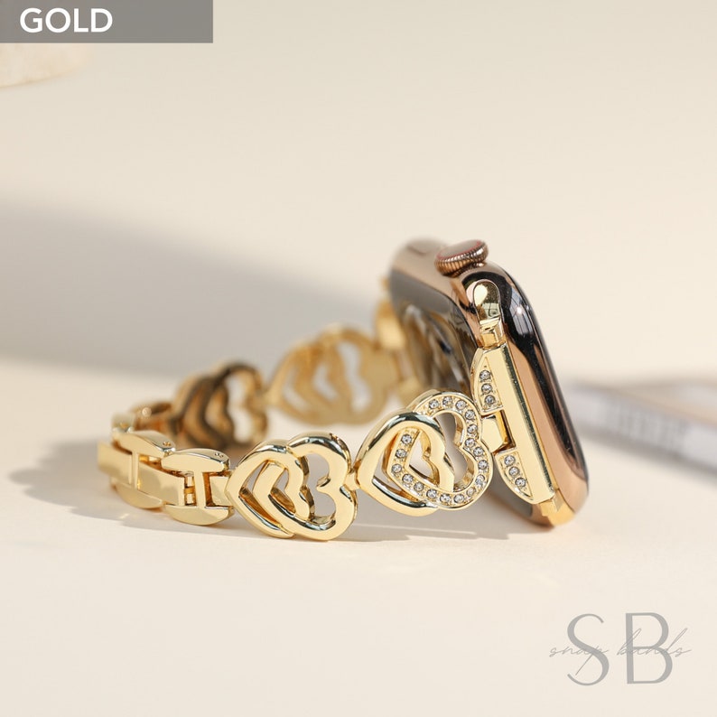 Gold Apple Watch band with a heart-shaped design and clear rhinestones. The band is made of metal and has a clasp closure.