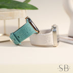 Two Apple Watch bands with floral designs. The band on the left is teal and the band on the right is white.