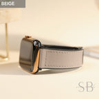 Beige Apple Watch with a light gray leather band. The watch has a black screen and a gold-colored frame. The band is stitched with a matching thread. The watch is a stylish accessory.