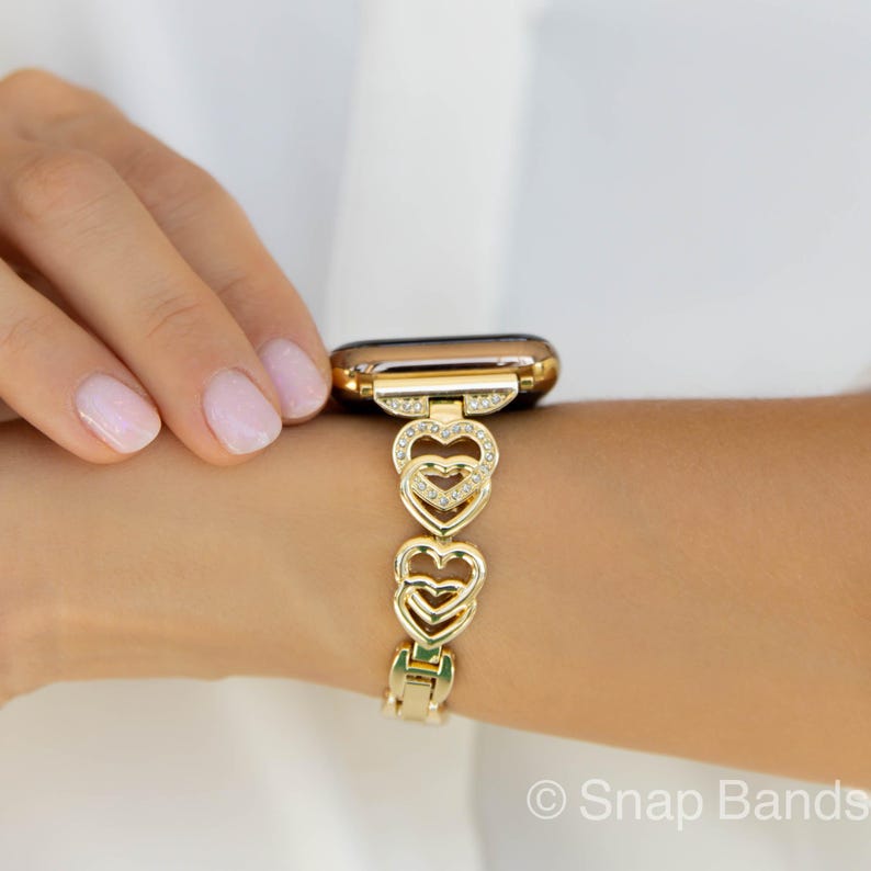Gold-tone Apple Watch band with a heart-shaped design and clear crystals. The band is made of metal and has a clasp closure.