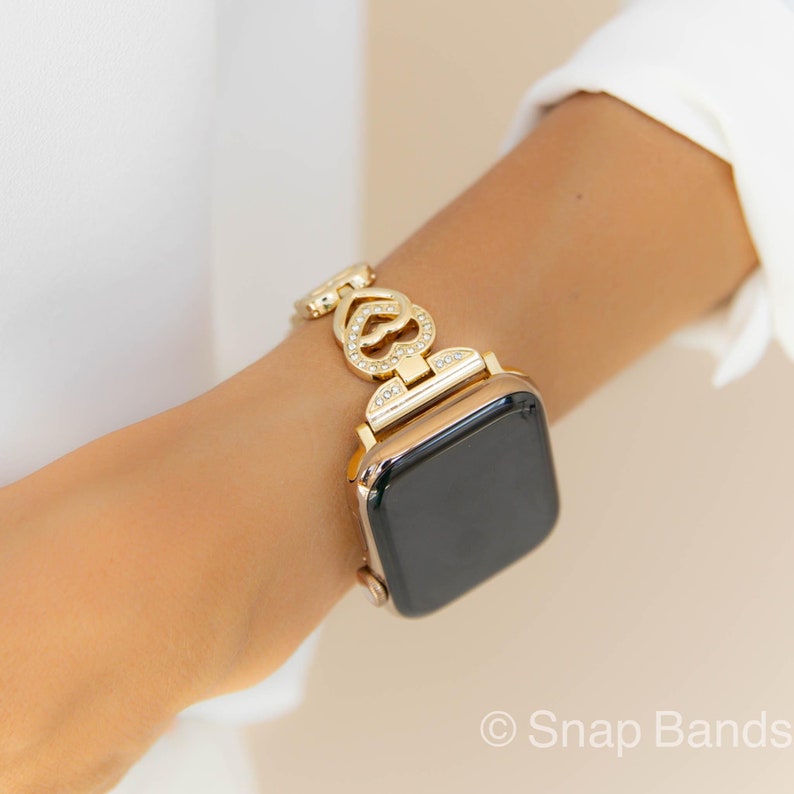 Gold Apple Watch band with a heart-shaped design and clear rhinestones. The band is made of metal and has a snap closure.