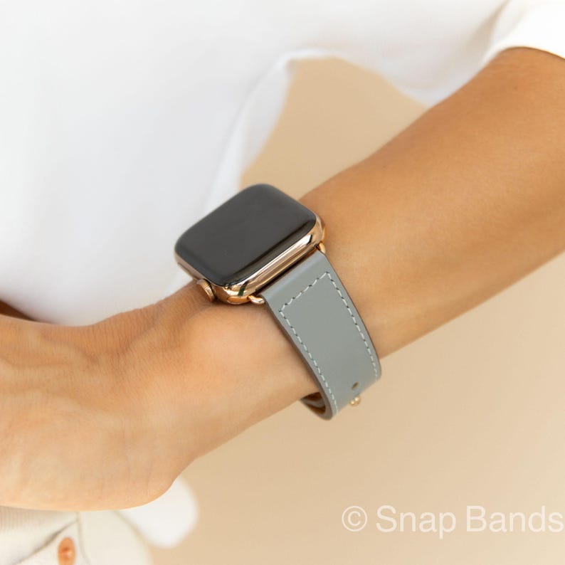 A gray leather watch band with a rose gold Apple Watch. The band has white stitching.
