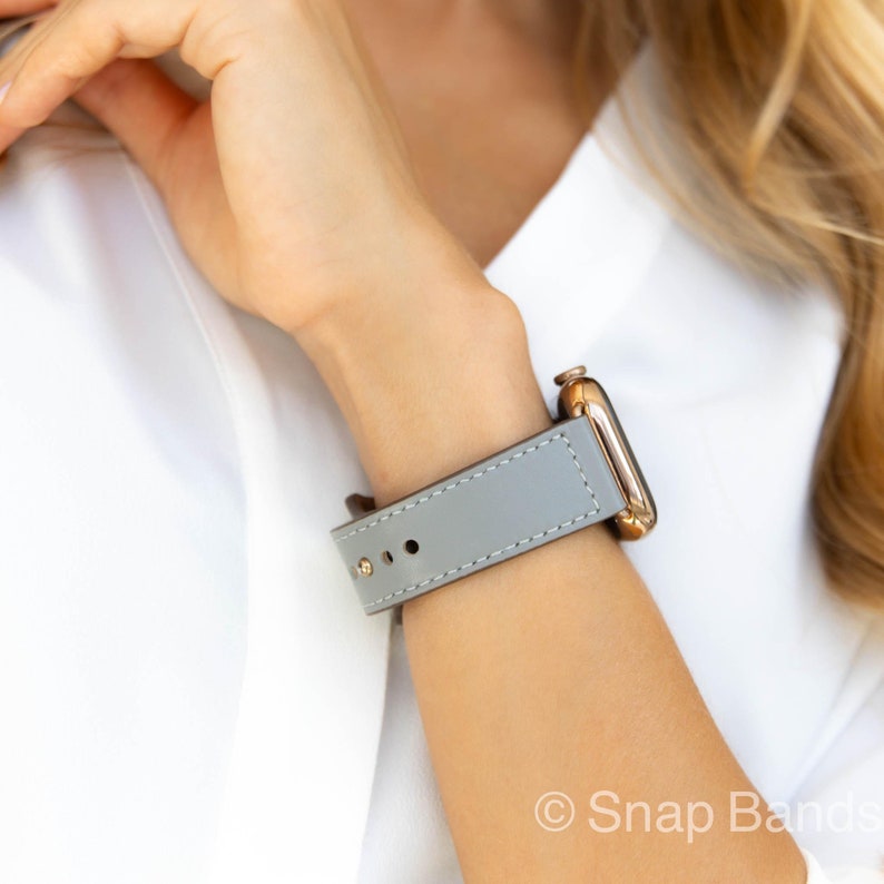 A gray leather watch band with a rose gold Apple Watch. The band has white stitching and is shown on a person's wrist.