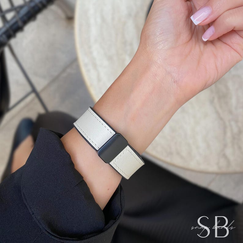 A white and black watch band is shown on a wrist. The watch band is made of white leather with black accents and stitching. The watch band is secured with a black clasp. The watch band is a stylish accessory.