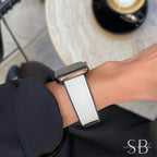 A rose gold smartwatch with a white leather band is displayed on a wrist. The band has a black underside and white stitching. The watch is worn against a blurred background.