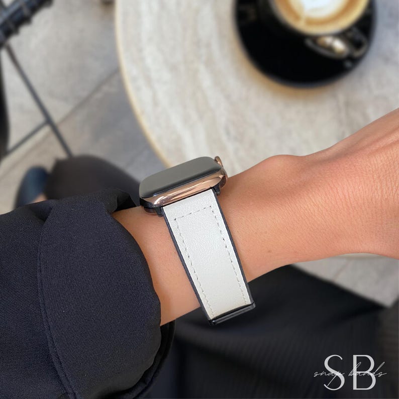A rose gold smartwatch with a white leather band is displayed on a wrist. The band has a black underside and white stitching. The watch is worn against a blurred background.