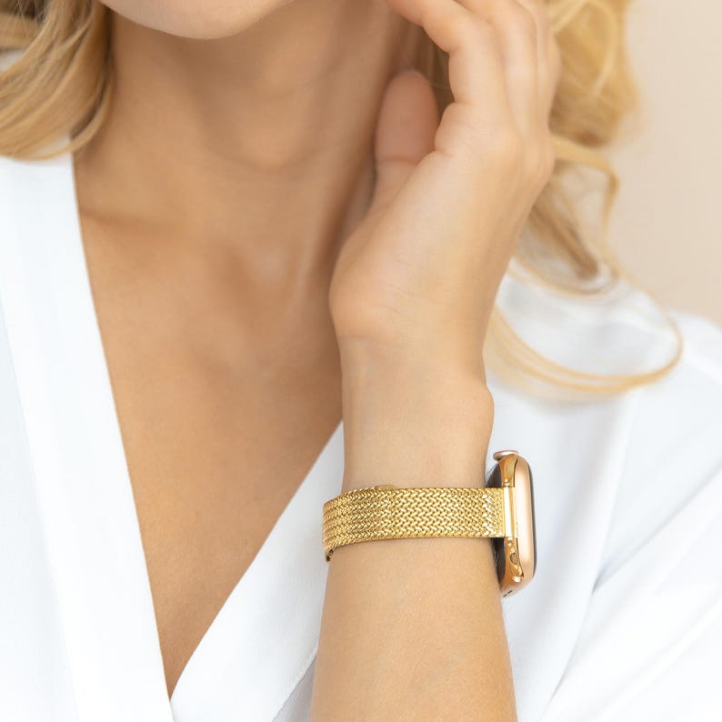 Gold-toned watch with a woven band. The watch case and band are a matching gold color. The band has a textured, interwoven design. The watch is secured on a wrist, with a white shirt in the background.