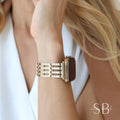 Gold-toned watch band with a rectangular design. The band is attached to a gold watch with a dark screen. The watch band has a series of horizontal bars. The watch is on a wrist.