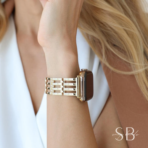 Gold-toned watch band with a rectangular design. The band is attached to a gold watch with a dark screen. The watch band has a series of horizontal bars. The watch is on a wrist.