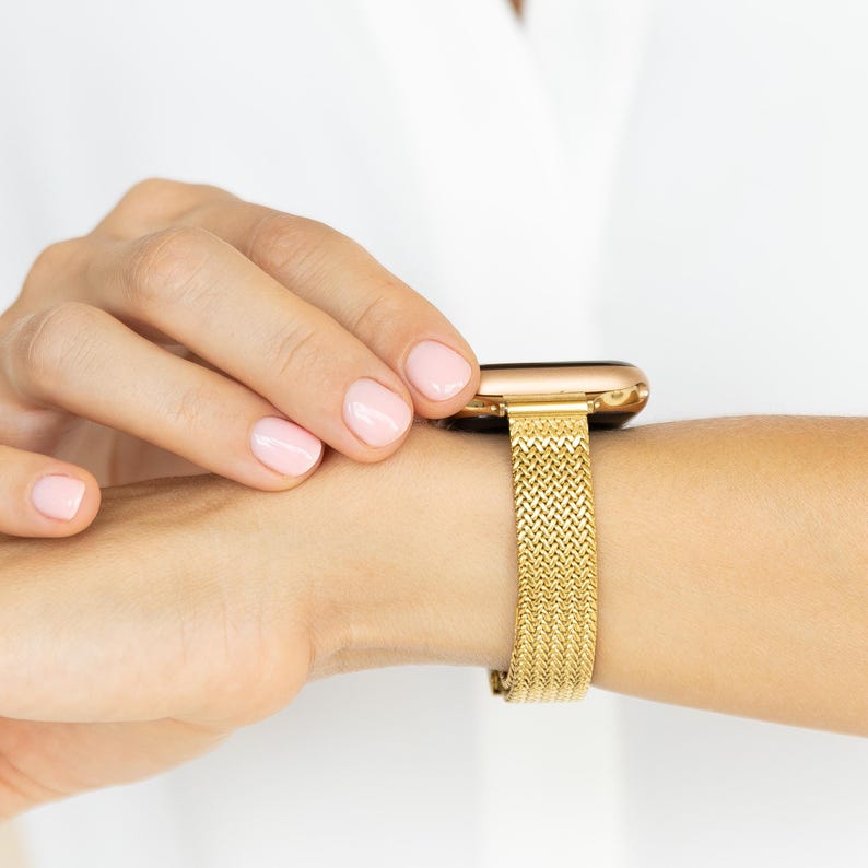 A gold-colored smartwatch with a matching gold mesh band is worn on a wrist. The band has a woven design. The person's hand is adjusting the watch. The background is white.