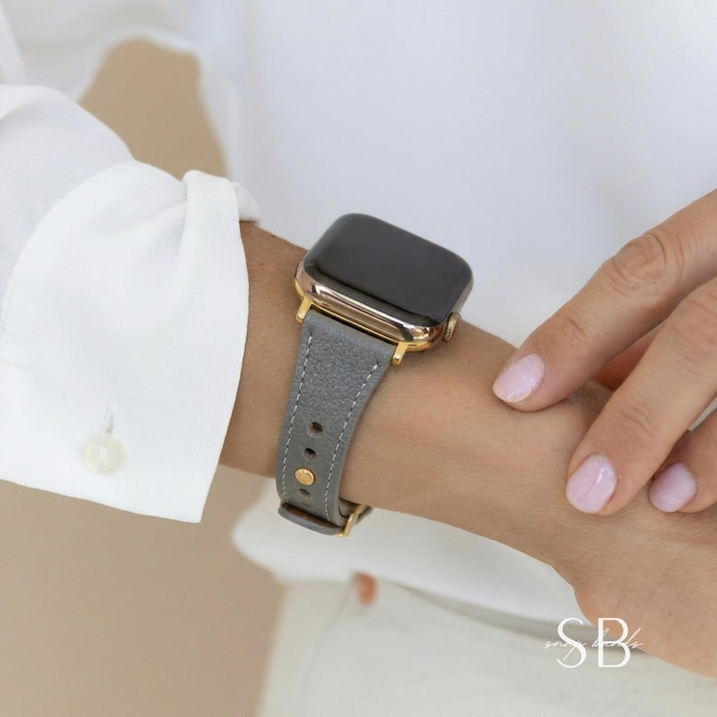 A gray leather watch band with gold-colored hardware is attached to a black smartwatch. The watch is worn on a wrist, with a white shirt sleeve and a hand with painted nails in the background.