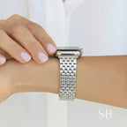 A silver-toned watch band with a rectangular link design is shown on a wrist. The watch face is square with a black screen. The band is made of metal and has a polished finish. The background is a white shirt.
