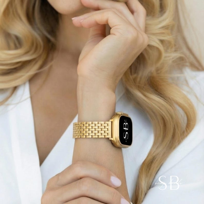 Gold-toned smartwatch with a rectangular face and a matching link band. The watch face is black with white text. The band is made of small, rectangular links. The initials "SB" are visible on the watch face.