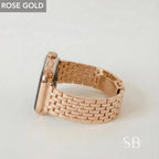Rose gold smartwatch with a matching band. The band is made of small, rectangular links. The watch face is black. The text "ROSE GOLD" is in a gray box in the upper left corner. The letters "SB" are in the lower right corner.
