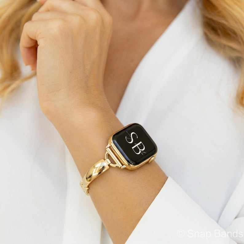 A gold-toned Apple Watch with a black screen displaying the initials "SB" in white. The watch is attached to a matching gold band with a woven design. The watch is worn on a wrist, against a white background.