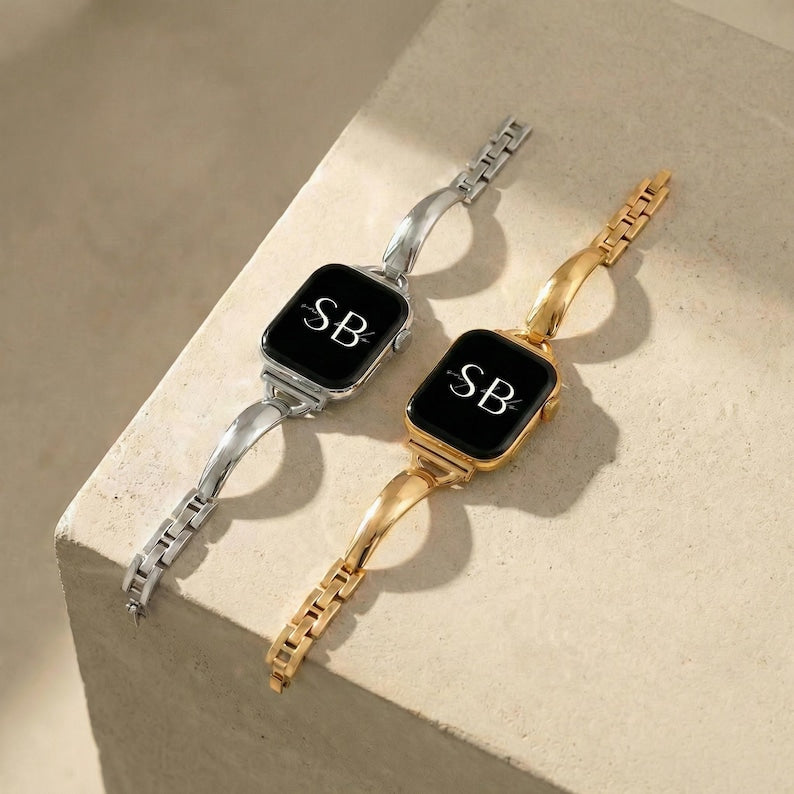 Two smartwatches with black screens, one silver and one gold. Each watch displays the initials "SB" in white. The bands are made of linked metal segments and curved, solid metal. The watches are displayed on a light-colored surface.