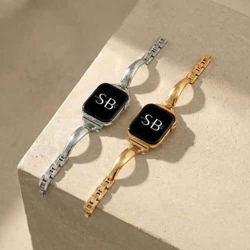 Two smartwatches with black screens, one silver and one gold. Each watch displays the initials "SB" in white. The bands are made of linked metal segments and curved, solid metal. The watches are displayed on a light-colored surface.