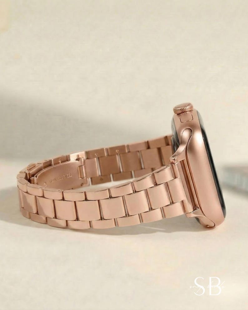 Rose gold smartwatch with a matching metal band. The watch face is black, and the band is made of rectangular links. The watch is shown from a side profile, displaying the crown and the band's clasp.