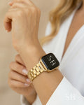 Gold-toned smartwatch with a matching metal band. The watch face is black with the initials "SB" in white. The band is made of rectangular links. The watch is worn on a wrist, with a white shirt in the background.