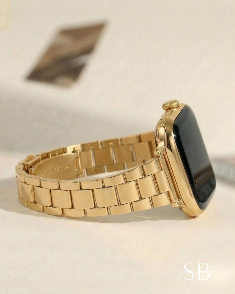 A gold-colored smartwatch with a matching metal band. The watch face is black, and the band is composed of rectangular links. The watch is shown from a side angle, highlighting its sleek design and metallic finish.
