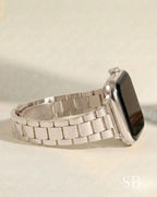 A silver-toned smartwatch with a rectangular face and a matching metal band. The band is composed of rectangular links. The watch is displayed on a light-colored surface.