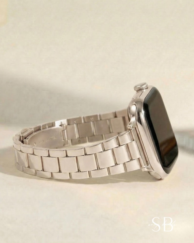 A silver-toned smartwatch with a rectangular face and a matching metal band. The band is composed of rectangular links. The watch is displayed on a light-colored surface.
